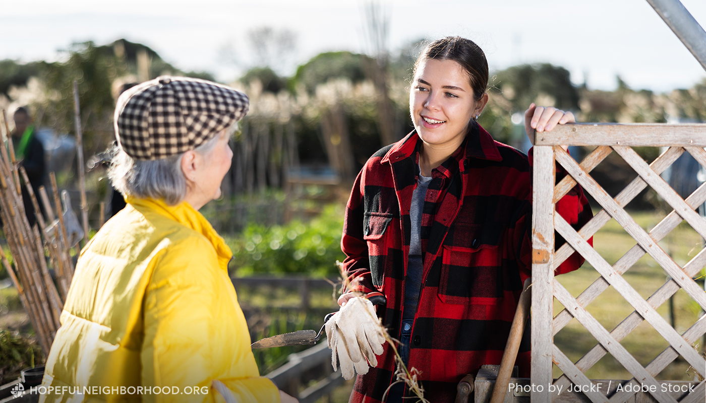 Older woman in a yellow winter coat and hat is talking to a younger woman in red and black flannel. The women are outside near a garden. This is an example of how you build trust in your community.