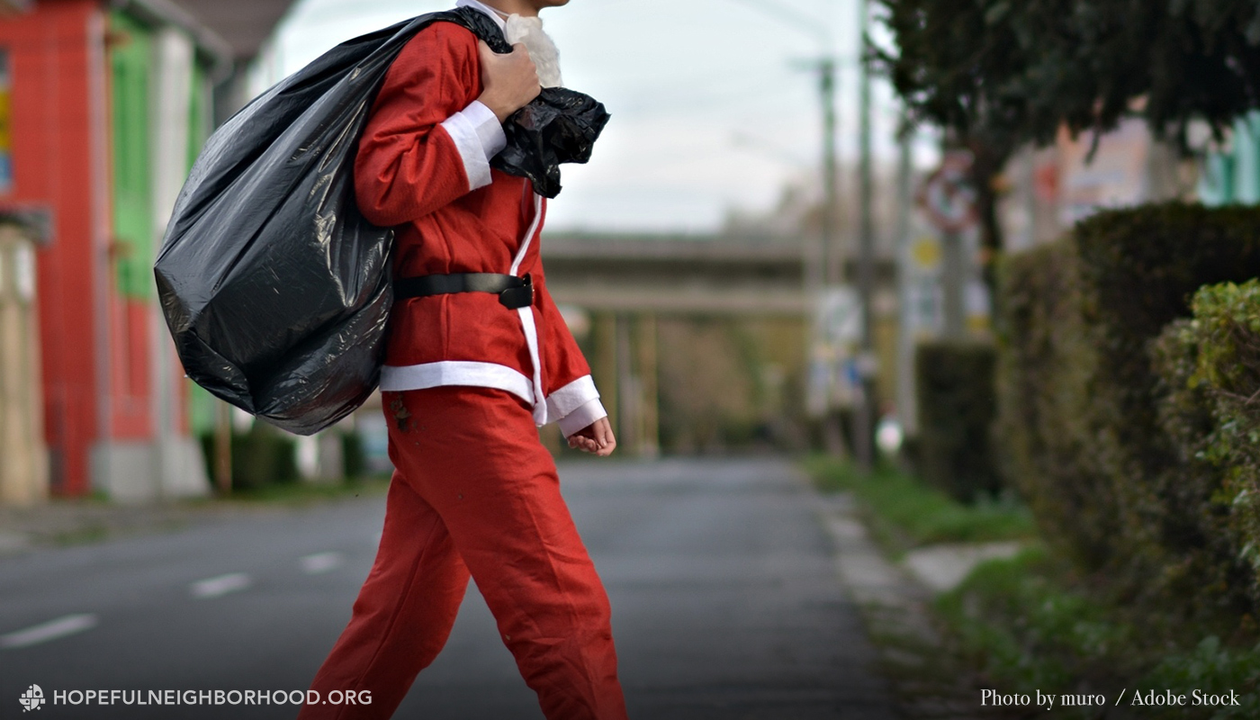 Photo of a skinny Santa carrying a black trash bag, crossing a street in an urban area.