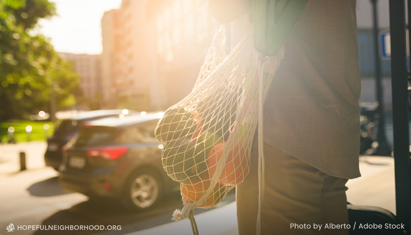 Image of a man along a street holding a reusable bag of produce. View of a city in background.