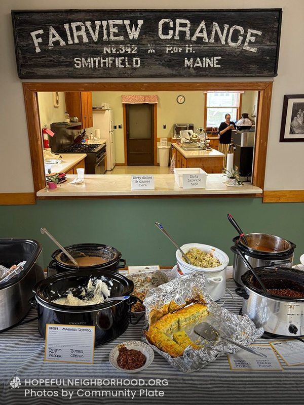 Photo shows an old sign with the text FAIRVIEW GRANGE Smithfield Maine in the background, with a table full of crockpots and recipe cards in the front.
